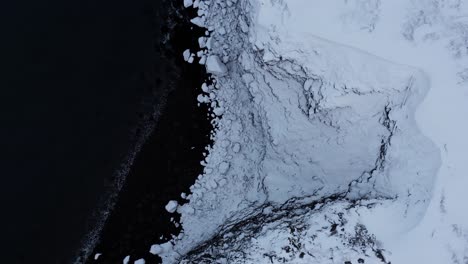 Schneebedeckte-Küstenklippe-In-Island-Mit-Schwarzem-Vulkanischem-Sandstrand,-Von-Oben-Nach-Unten-Antenne