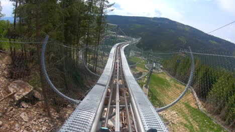 POV-GoPro-shot-of-a-man-riding-on-an-elevated-fenced-outdoor-roller-coaster-in-the-mountains-of-Dolní-Morava,-Czech-Republic,-descending-down-in-a-spiralling-loop