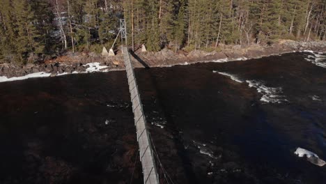 Reveal-aerial-shot-of-a-suspension-bridge-in-Mårdseleforsen,-crossing-the-Vindelälven-river,-located-in-the-north-of-Sweden,-unveiling-a-beautiful-riverbed-with-high-lush-century-old-pine-trees