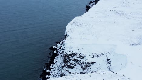 White-snow-covered-cliffs-along-calm-ocean-water,-Iceland,-aerial