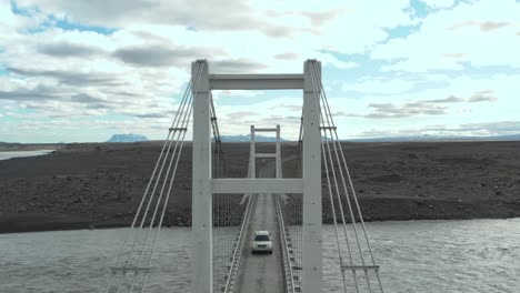 Car-crossing-on-single-lane-bridge-above-glacial-river-in-Iceland,-fly-through-steel-tower,-aerial