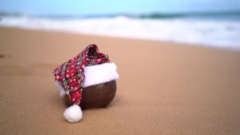 Close-up-of-a-coconut-wearing-santa-hat-on-tropical-sandy-beach-with-ocean-waves-rolling-against-coastline-in-blurred-background