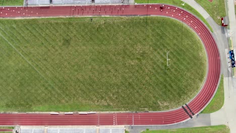 Top-down-aerial-birds-eye-view-of-red-track,-green-athletic-field,-student-athletes-practicing