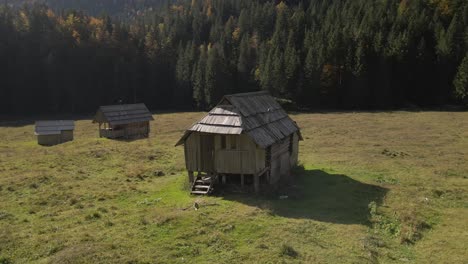 Old-wooden-cabin-in-grass-field-open-meadow-in-mountains,-aerial