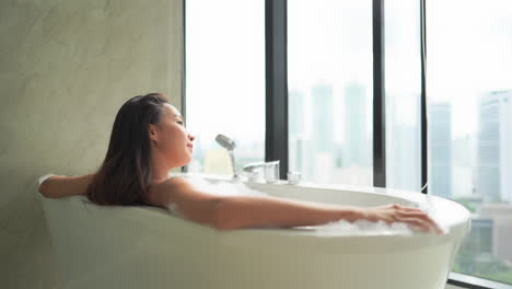 While-relaxing-in-a-large-resort-style-bathtub,-a-young-beautiful-woman-looks-out-on-a-modern-skyline