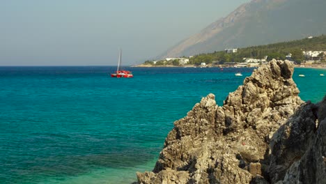 Mediterranean-coastline-with-rocks-and-blue-turquoise-sea,-anchored-boats-near-touristic-village-in-Albania