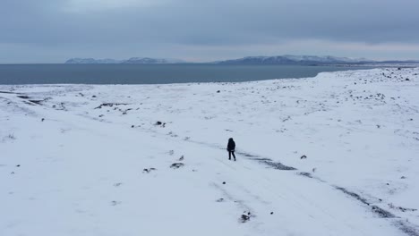 Man-walking-towards-coastal-cliffs-in-white-snow-covered-landscape,-aerial