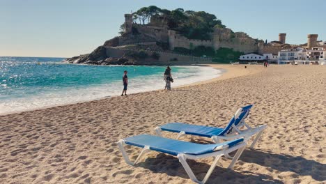 Deck-chairs-on-the-beach-with-the-background-of-walled-castle-over-the-sea-in-Tossa-de-Mar,-Girona-Spain-Costa-Brava-turquoise-water-beaches