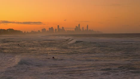 Meereswellen,-Die-Mit-Surfern-Am-Strand-Rollen---Orangefarbener-Sonnenuntergang-Mit-Skylinen-Des-Surferparadieses---Goldküste,-Queensland,-Australien