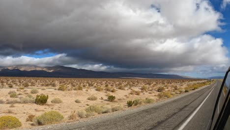 View-of-the-Mojave-Desert-landscape-with-dark,-ominous-clouds-overhead-as-seen-from-a-vehicle-driving-through-and-looking-back