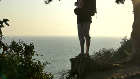 Young-man-wearing-a-large-travel-backpack-standing-in-silhouette-on-a-tree-stump-looking-out-over-a-Pacific-ocean-cliff-during-a-golden-sunset-in-Santa-Barbara,-California