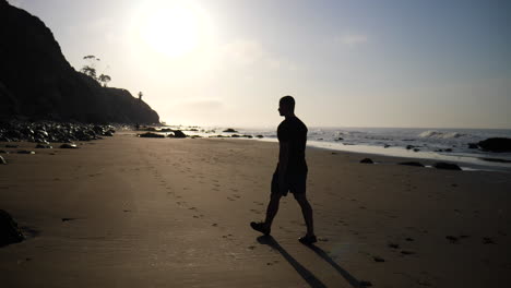 A-man-in-silhouette-walking-on-the-beach-at-sunrise-with-ocean-waves-crashing-on-the-coast-in-Santa-Barbara,-California-SLOW-MOTION