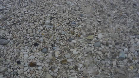 Pebbles-on-shore-wallow-by-clear-crystal-water-of-sea-waves,-seabed-background-for-copy-space