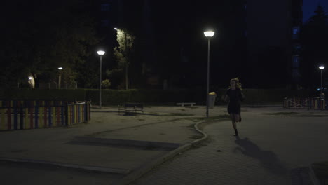 Scared-Woman-In-Black-Casual-Dress-Running-Away-Passing-By-An-Empty-Park-With-Street-Lamps-Late-At-Night-In-Spain
