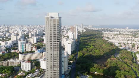 Scenic-city-skyline-aerial-view-of-tall-residential-Avenida-Anacaona-skyscraper-and-downtown-Santo-Domingo-center-and-view-of-ocean-sea-water-on-sunny-day,-Dominican-Republic,-overhead-drone-descend
