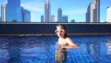 A-young-woman-in-a-private-rooftop-resort-pool-turns-from-side-to-side-against-an-urban-skyline