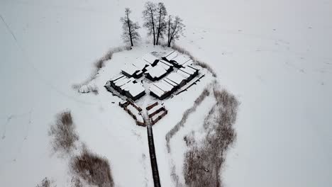 Snow-covered-wooden-buildings-surrounded-by-white-frozen-lake,-aerial-drone-view