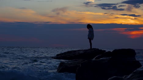 Young-woman-standing-on-cliff-splashed-by-sea-waves-at-beautiful-sunset-background-of-cloudy-sky