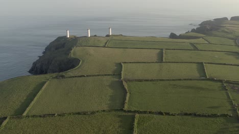 Rising-evening-coastal-farm-aerial-of-Metal-Man-beacon-towers,-IRL