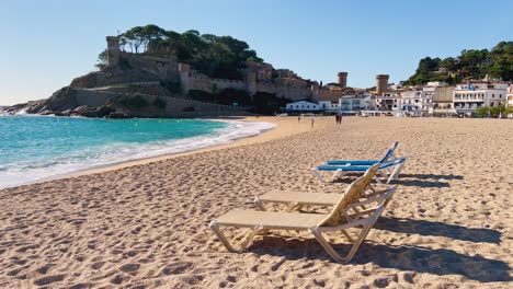 Liegestühle-Am-Strand-Mit-Dem-Hintergrund-Der-Ummauerten-Burg-über-Dem-Meer-In-Tossa-De-Mar,-Girona,-Spanien,-Costa-Brava,-Türkisfarbene-Wasserstrände