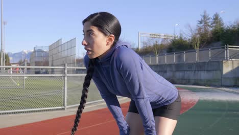 Runner-woman-stops-to-catch-breath-with-hands-on-knees-on-race-track,-Handheld