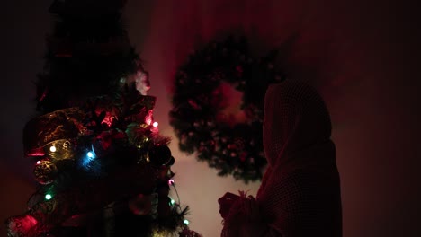 Woman-In-The-House-Wearing-Scarf,-Hangs-Ornaments-On-The-Christmas-Tree-With-Colorful-Dim-Lights