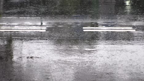 Raindrops-causing-ripples,-falling-on-reflective-black-tarmac-road-with-white-road-markers