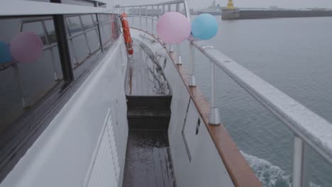 Walking-along-starboard-side-of-yacht-on-rainy-day,-traveling-towards-harbor,-pink-and-blue-balloon,-luxury-gender-reveal-party