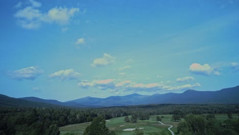 The-white-mountain-range-in-New-Hampshire-sits-under-a-blue-cloudy-sky-beyond-forest,-which-surrounds-a-gold-course
