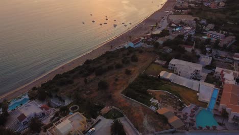 Aerial-shot-of-orange-sunset,-beach-full-od-umbrellas,-hotels-and-small-floating-boats-in-Greece