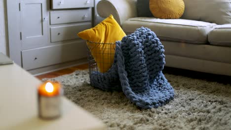 Metal-Basket-With-Pillow-And-Warm-Hand-Knit-Blanket,-Cozy-Living-Room-With-Lit-Candle-On-Table---static-shot