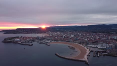 Aerial-panoramic-view-of-gijon-cityscape-during-epic-sunset,-beach-urban-city-center-building-and-harbor-with-mountains-in-background