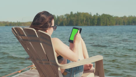 Young-woman-relaxes-on-a-adirondack-chair-on-a-lakeside-dock-in-the-summer-with-a-green-screen-for-chroma-key