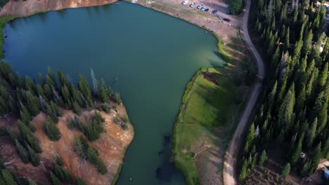 Beautiful-aerial-drone-shot-of-a-stunning-nature-landscape-of-the-Anderson-Meadow-Reservoir-lake-up-Beaver-Canyon-in-Utah-with-large-pine-tree-forest,-a-dirt-road-and-a-parking-lot-for-fisherman