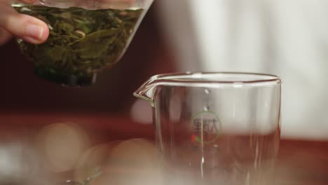 Pouring-infused-green-tea-from-gaiwan-bowl-into-the-glass-on-traditional-dark-wood-tea-table