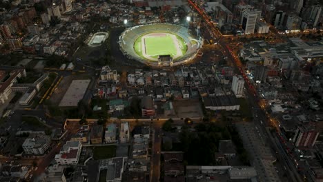 Toma-Aérea-Inclinada-Que-Muestra-El-Paisaje-Urbano-De-La-Ciudad-De-Quito-En-Ecuador-Con-Estadio-De-Fútbol-Iluminado-Durante-El-Juego-Y-Nubes-Místicas-Entre-Montañas-En-Segundo-Plano