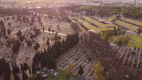 Aerial-flyover-large-Chacarita-Cemetery-at-sunset-with-sun-flares-in-Buenos-Aires---Trees-and-path-between-graves