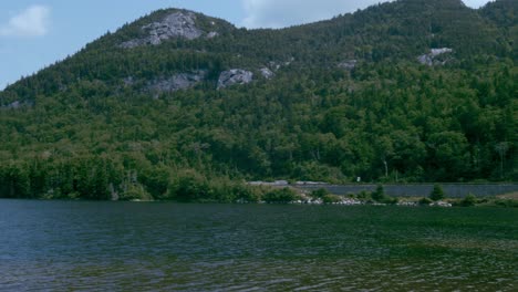 Ein-Mit-Vegetation-Bedeckter-Granitberg-Steht-über-Einem-Ruhigen-See-Vor-Blauem-Himmel,-Während-Die-Kamera-Schwenkt