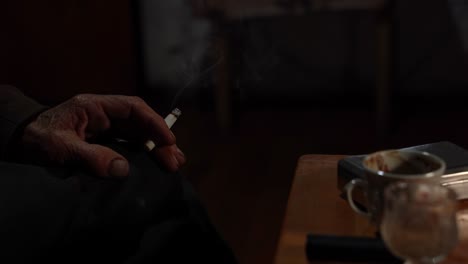 Old-man-hand-holding-smoking-cigarette-near-coffee-cup-in-a-dark-room,-elderly