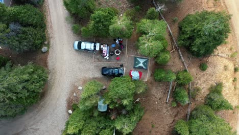 Bird's-eye-aerial-top-view-of-a-small-campground-with-tents,-cars,-a-fire-pit,-and-large-green-pine-trees-surrounding-them-on-a-warm-sunny-summer-morning-in-Anderson-Meadow-Reservoir-in-Beaver,-Utah