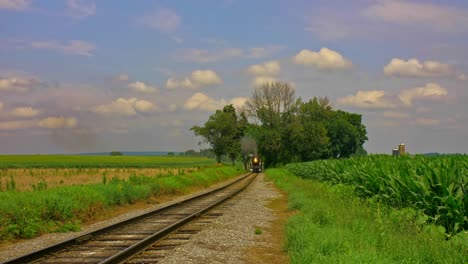 A-View-of-a-Steam-Passenger-Train-Approaching-Thru-the-Trees-Blowing-Black-Smoke-by-Corn-Fields-on-a-Partially-Cloudy-Summer-Day