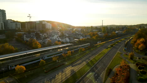 Passenger-Railway-train-arriving-at-Gdynia-Redlowo-station-early-in-the-morning-at-bright-Autumn-sunrise-aerial-view,-several-cars-moving-on-droga-Gdynska-highway