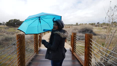 Eine-Hübsche-Frau,-Die-Während-Eines-Regensturms-Bei-Schlechtem-Wetter-In-Zeitlupe-über-Eine-Brücke-Mit-Einem-Blauen-Regenschirm-Geht