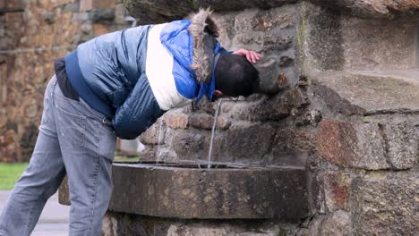Young-man-wearing-wide-jacket-drinks-from-the-water-spout-of-a-stone-fountain-on-a-rural,-small-village