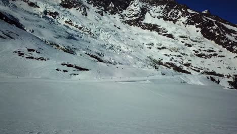 Vista-De-Drones-De-Una-Llanura-De-Nieve-En-Los-Alpes-Suizos,-Telesilla-Y-Esquiadores-En-El-Fondo