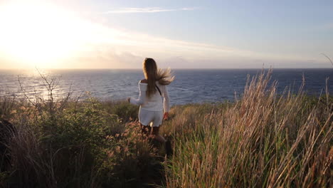 A-woman-wearing-white-walks-through-tall-grass-overlooking-a-beautiful-ocean-view-at-sunset