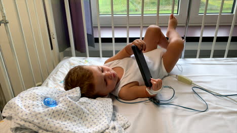 Young-baby-boy-laying-down-in-hospital-bed-watching-movies-on-smartphone