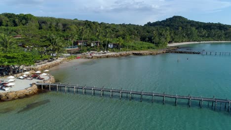 Aerial,-pan,-drone-shot-of-buildings-on-the-coast-and-wooden-piers,-in-turquoise-sea,-at-a-paradise-beach,-on-a-sunny-day,-in-Koh-Kood,-Thailand