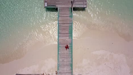 Aerial-perspective-straight-down-on-a-wooden-pier-in-Thailand,-then-tracking-man-as-he-runs-and-dives-into-clear-green-ocean-water,-Lookdown,-follow,-tracking,-4k