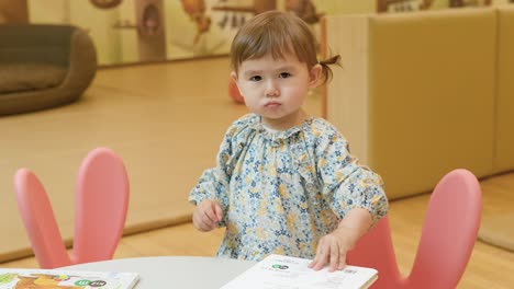 Adorable-2-year-old-girl-sits-at-the-table-and-looks-at-colorful-book-with-pictures-and-closing-book,-stand-up-and-look-at-camera-with-wise-frawn-face